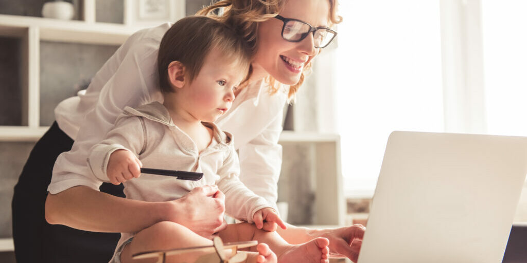 Beautiful business mom is using a laptop and smiling while spending time with her cute baby boy at home